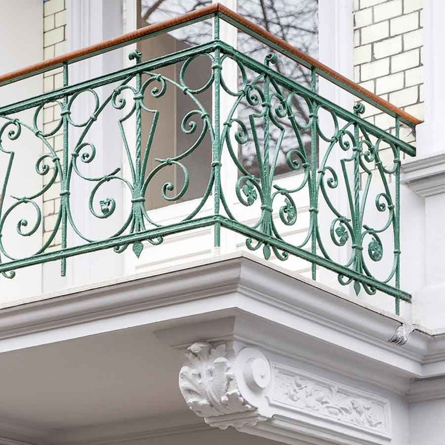 Detailaufnahme eines sanierten Altbau-Balkons mit einem filigranen, grünen schmiedeeisernen Geländer und Stuckdetails an der weißen Fassade Detailaufnahme eines sanierten Altbau-Balkons mit einem filigranen, grünen schmiedeeisernen Geländer und Stuckdetails an der weißen Fassade